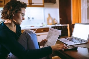 A student sits at her kitchen table with bills and a calculator, laptop open in front of her