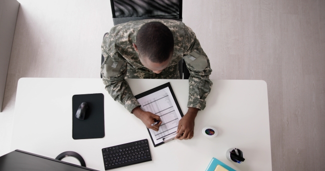 Aerial shot of a man in a U.S. military uniform sitting at a white desk, filling out paperwork.