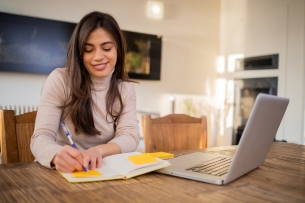 A smiling woman in front of a laptop writes in a notebook. 