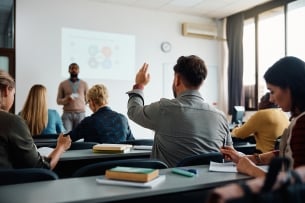 Back view of student raising his hand to answer a teacher’s question in class while other students look down and don’t have their hands raised.