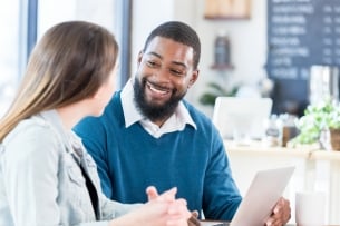 An African American man sits in a coffee shop with a woman and uses his laptop during their discussion.