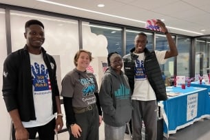 Four students stand in front of a table with a table banner that reads “Cuyahoga Community College.” Two wear shirts with the slogan “Your voice matters. #voiceyourvote.”