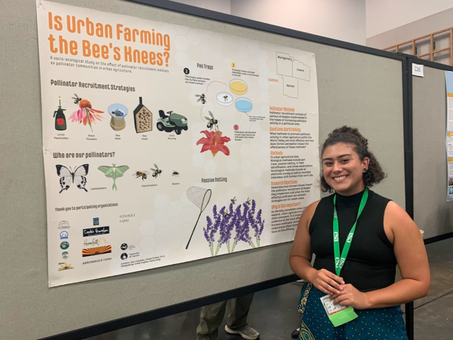 A biology student stands in front of a poster featuring information on bees and other pollinators